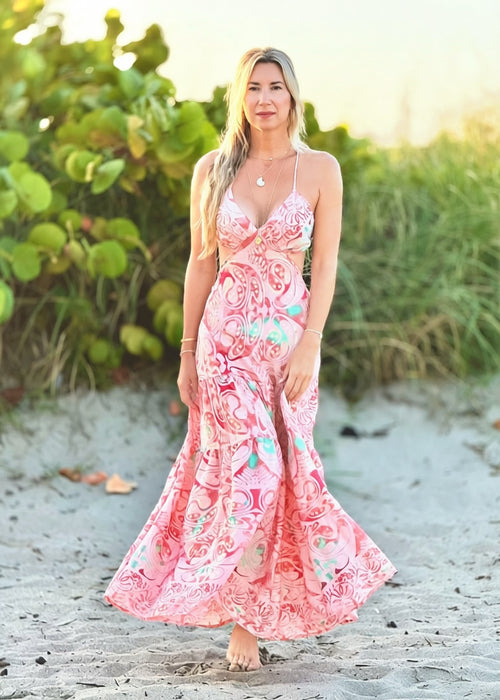 Woman in a pink floral dress standing on a sandy beach with greenery in the background