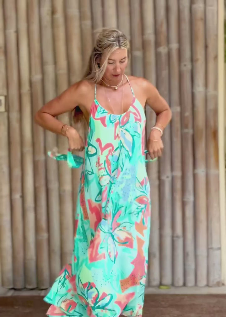 A woman modeling a sleeveless maxi dress with a vibrant tropical floral print in turquoise, coral pink, and green, standing barefoot in front of a bamboo wall.