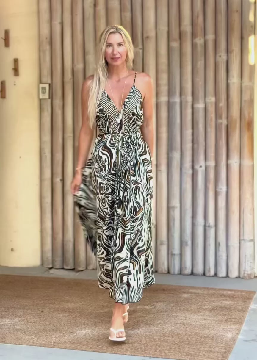 A woman modeling a sleeveless maxi dress with a black, white, and gray abstract swirl pattern, standing in front of a bamboo wall.
