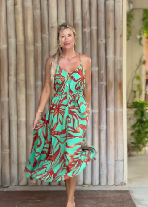 A woman wearing a sleeveless midi dress with a vibrant turquoise, orange, and red abstract pattern, standing barefoot in front of a bamboo wall.