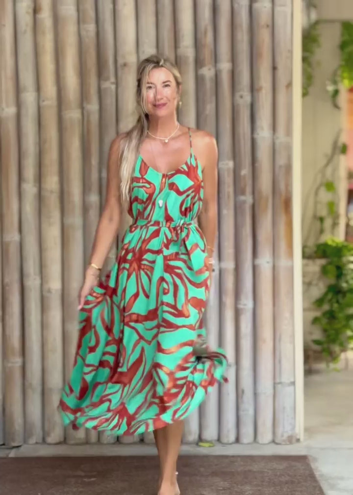 A woman wearing a sleeveless midi dress with a vibrant turquoise, orange, and red abstract pattern, standing barefoot in front of a bamboo wall.