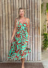 A woman wearing a sleeveless midi dress with a vibrant turquoise, orange, and red abstract pattern, standing barefoot in front of a bamboo wall.