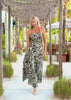 A woman modeling a sleeveless halter-neck maxi dress with a black, white, and brown abstract swirl pattern, walking on a patio under a rustic wooden pergola.