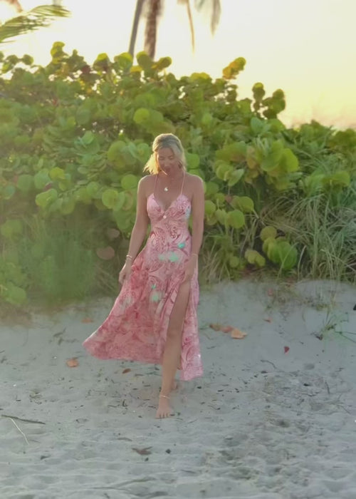 Woman in a pink floral dress standing on a beach with greenery in the background