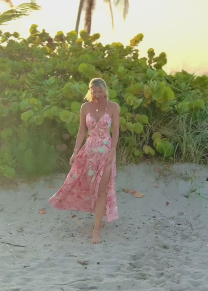 Woman in a pink floral dress standing on a beach with greenery in the background
